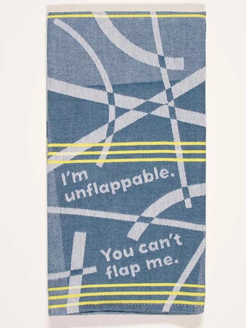 A blue denim-style woven Blue Q tea towel with yellow pinstripes and geometric intersecting white lines, printed with "I'm unflappable. You can't flap me."