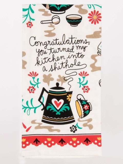 A white folded Blue Q tea towel with folk art floral and teapot illustrations, spill stains, and script text reading "Congratulations, you turned my kitchen into a shithole".