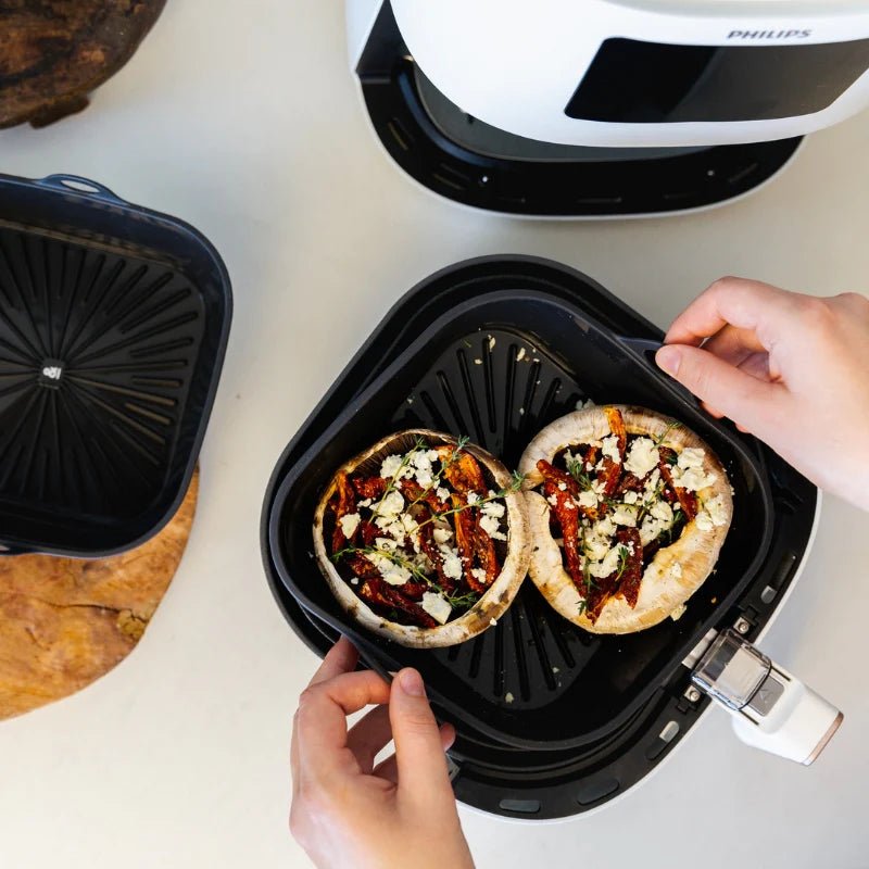 Hands lifting a liquorice black Seed & Sprout silicone air fryer bowl filled with stuffed mushrooms out of an air fryer basket.