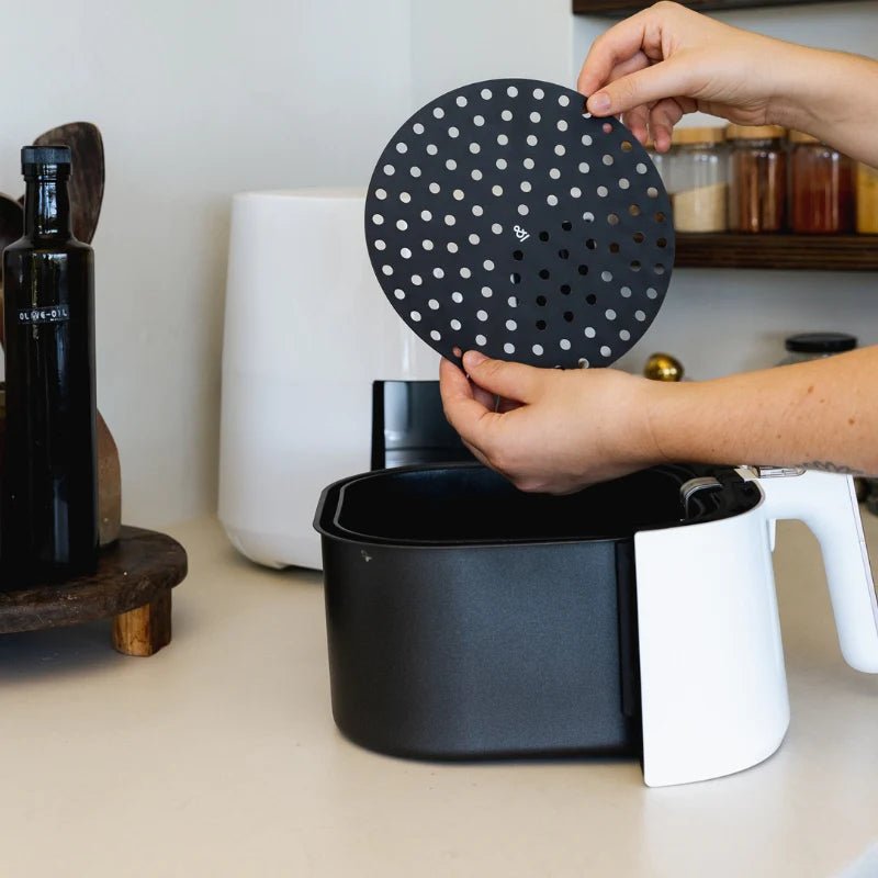 A pair of hands placing a black, perforated Seed & Sprout silicone air fryer mat into the basket of a modern white air fryer on a kitchen counter.