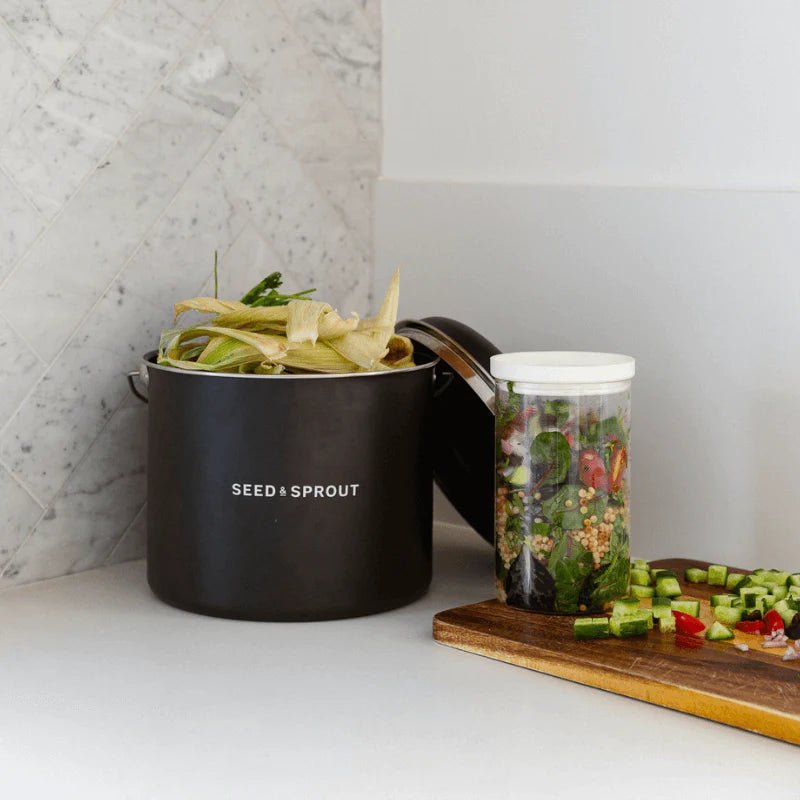Seed & Sprout black liquorice kitchen compost bin filled with organic food scraps sitting on a countertop next to chopped vegetables and a jar of salad.