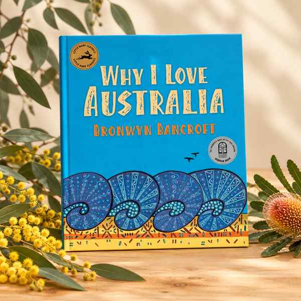 Why I Love Australia children's book by Bronwyn Bancroft with a blue cover, displayed on a wooden surface alongside native Australian wattle blossoms, eucalyptus leaves, and a banksia flower.