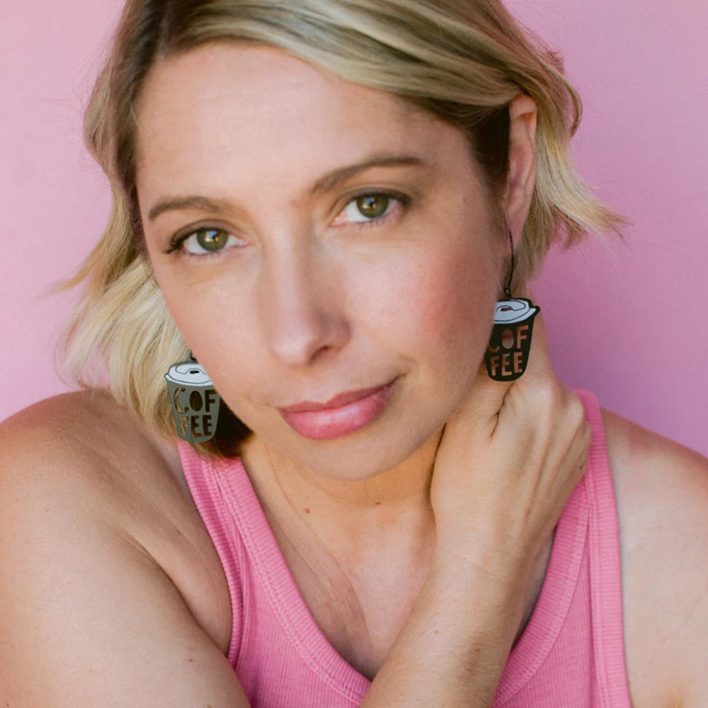 A close-up portrait of a woman with blonde hair wearing DDENZ Coffee Cup earrings with the text 'COFFEE' on them, posed against a solid pink background.