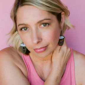 A close-up portrait of a woman with blonde hair wearing DDENZ Coffee Cup earrings with the text 'COFFEE' on them, posed against a solid pink background.