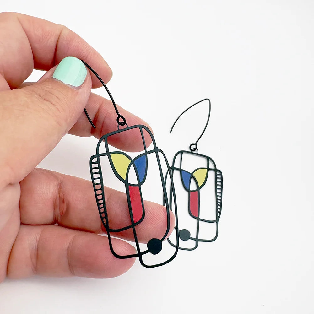 Hand holding two DENZ Art School Drop Out Earrings in Primary with colorful design on a white background