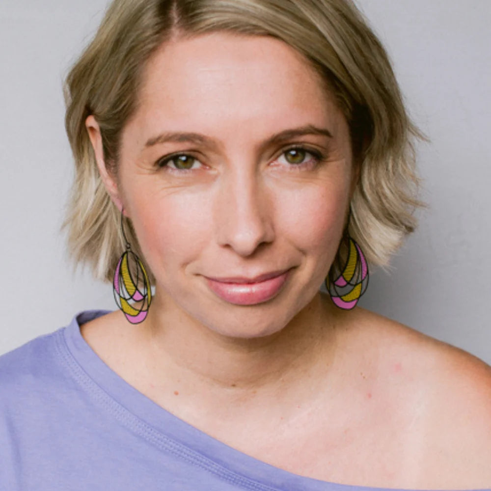 A close-up portrait of a woman with short blonde hair wearing DArt School Drop Out earrings and a lavender off-the-shoulder top against a plain light gray background.