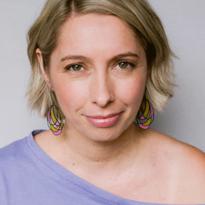 A close-up portrait of a woman with short blonde hair wearing DArt School Drop Out earrings and a lavender off-the-shoulder top against a plain light gray background.