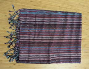 A close-up, top-down photograph of a woven textile featuring horizontal stripes in shades of aubergine, berry, and teal, finished with a twisted fringe along the left edge and displayed on a wooden surface.