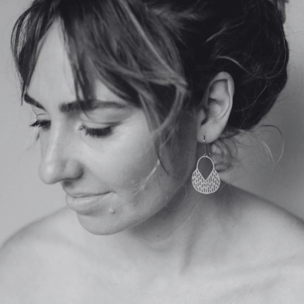 A close-up black and white portrait of a woman wearing a DENZ Callisia gold dangling geometric earring, with her face in profile and the earring clearly visible against a soft neutral background.