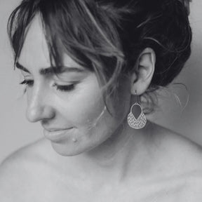 A close-up black and white portrait of a woman wearing a DENZ Callisia gold dangling geometric earring, with her face in profile and the earring clearly visible against a soft neutral background.