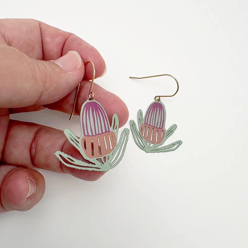 A pair of DENZ dangling earrings shaped like banksia flowers, shown with one earring held in a person's hand and the other hanging beside it on a plain white background.