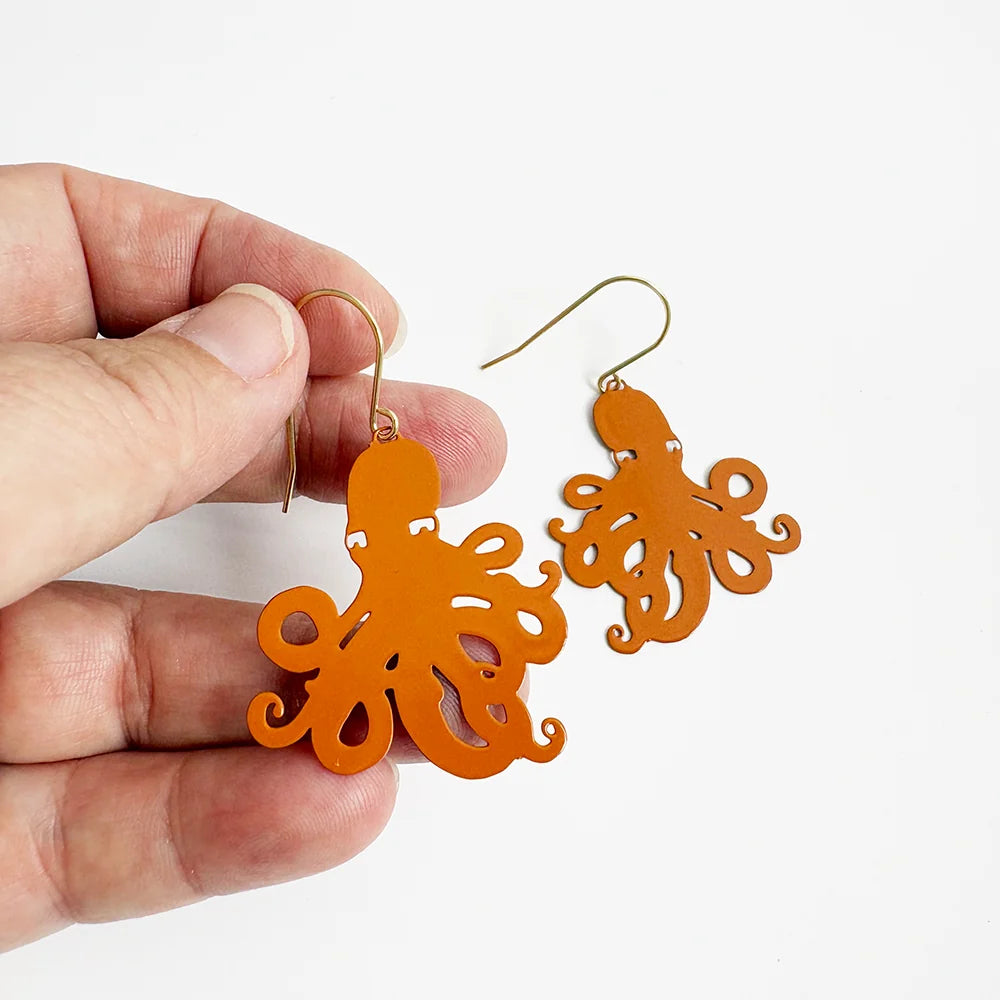 Orange DENZ octopus-shaped dangle earrings with gold-tone fishhook wires, shown with one held in a hand against a plain white background.