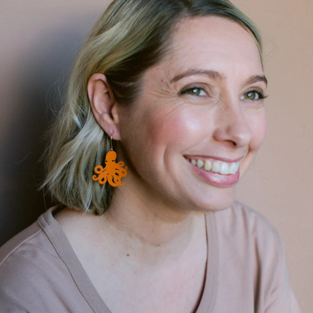 Woman wearing DENZ orange octopus-shaped earrings, smiling against a neutral background