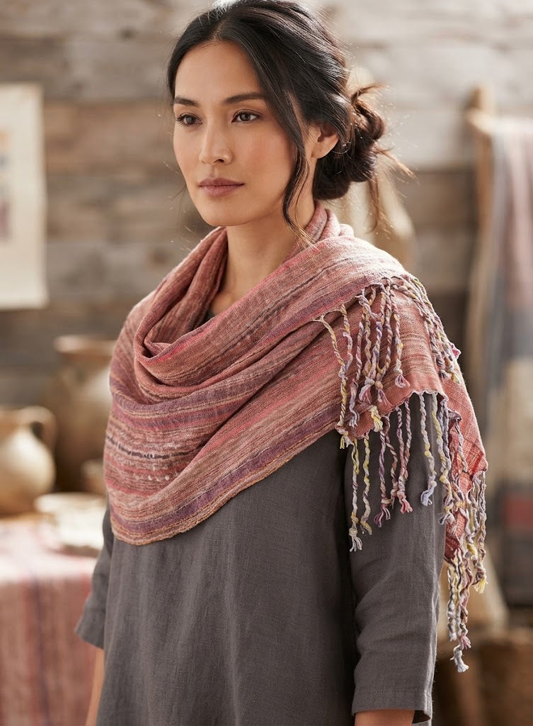 A close-up studio photograph of a woven shawl draped stylishly over the shoulders of a woman. The textile features a delicate vertical striped pattern in soft shades of dusty rose, mauve, plum, and subtle hints of gold and cream, creating a warm and textured look. The shawl is finished with intricate, multicolored knotted fringe tassels hanging neatly along the edge.