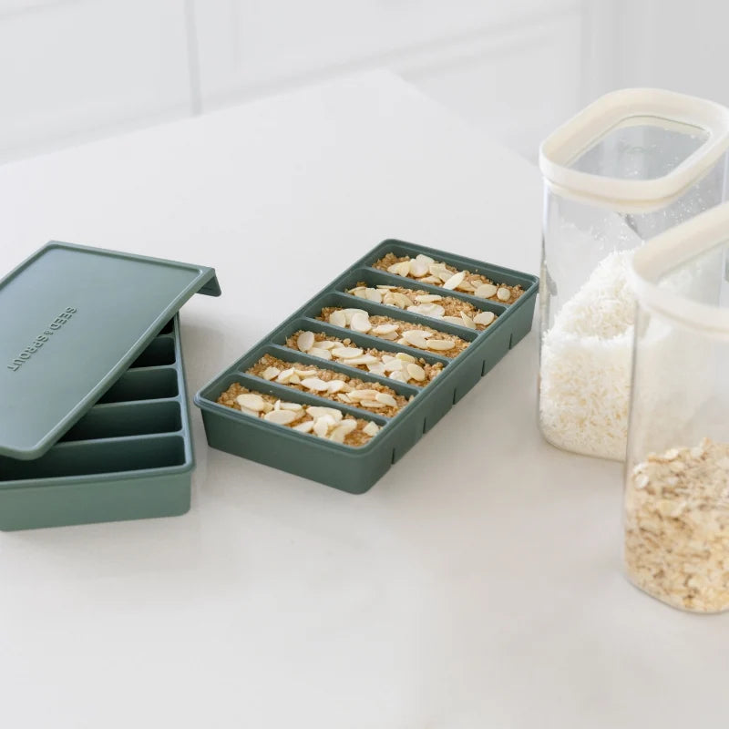 Seed & Sprout 6-portion silicone freezer tray in macadamia, featuring six 100ml compartments filled with almond-topped oat bars alongside an airtight lid and pantry containers.
