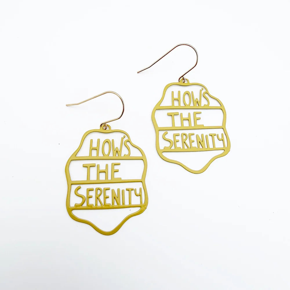 A pair of DENZ gold-tone dangle earrings with a scalloped outline and the text 'HOWS THE SERENITY' cut out, shown on a plain white background.