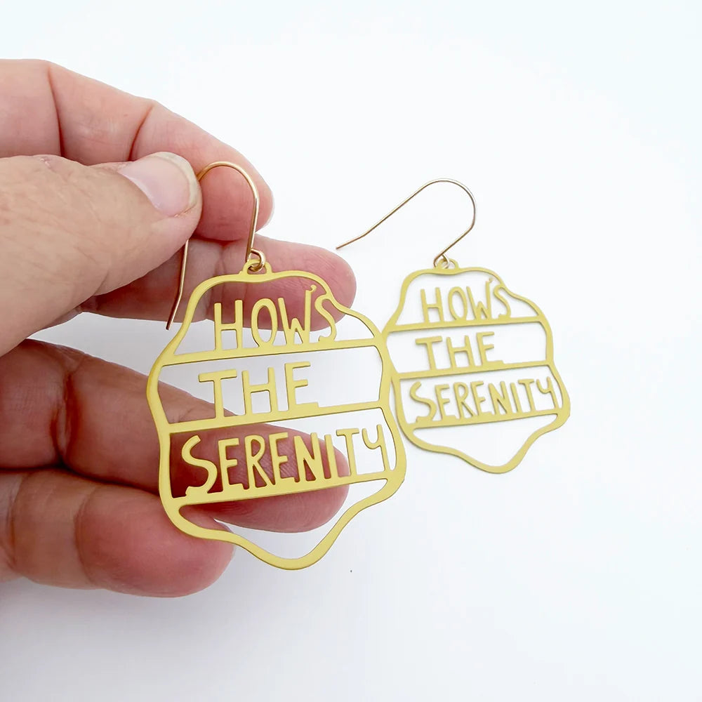 A hand holding a pair of DENZ gold-tone statement earrings with the text 'HOW'S THE SERENITY' in a cutout design, against a plain white background.