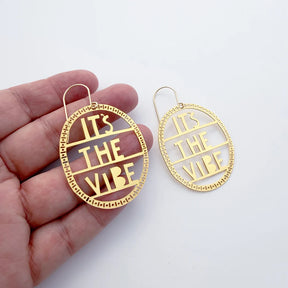 A pair of DENZ gold-toned oval drop earrings with cutout text reading 'IT'S THE VIBE' are shown, with one earring held in a person's hand and the other displayed beside it on a plain white background.
