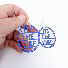 DENZ Indigo 'IT'S THE VIBE' earrings held by a hand against a white background