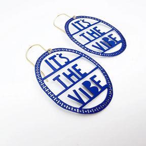 A pair of DENZ blue oval statement earrings with gold-tone ear wires, featuring the text 'IT'S THE VIBE' in a cutout design, set against a plain white background.