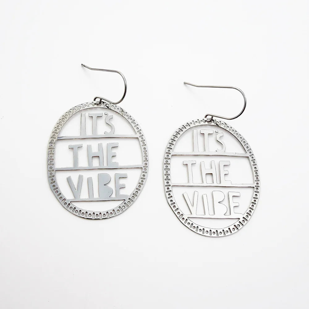 A pair of DENZ silver-toned oval dangle earrings with cutout text reading 'IT'S THE VIBE' and a beaded circular border, shown on a plain white background.