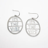 A pair of DENZ silver-toned oval dangle earrings with cutout text reading 'IT'S THE VIBE' and a beaded circular border, shown on a plain white background.