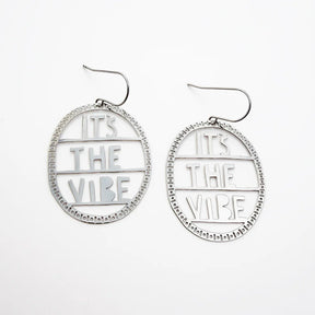 A pair of DENZ silver-toned oval dangle earrings with cutout text reading 'IT'S THE VIBE' and a beaded circular border, shown on a plain white background.