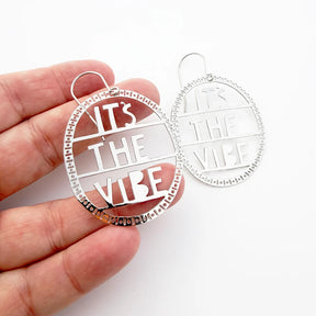 A hand holds a pair of DENZ silver-toned oval earrings with cutout text reading 'IT'S THE VIBE' on a plain white background.