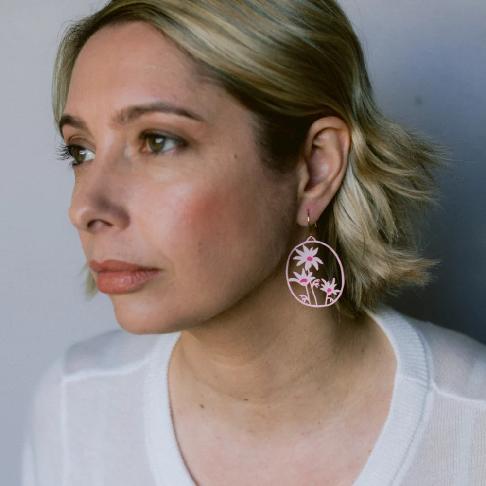 A close-up side profile of a woman with short blonde hair wearing a DENZ Flannel Flower earring, paired with a white knit top against a soft gray background.