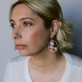 A close-up side profile of a woman with short blonde hair wearing a DENZ Flannel Flower earring, paired with a white knit top against a soft gray background.