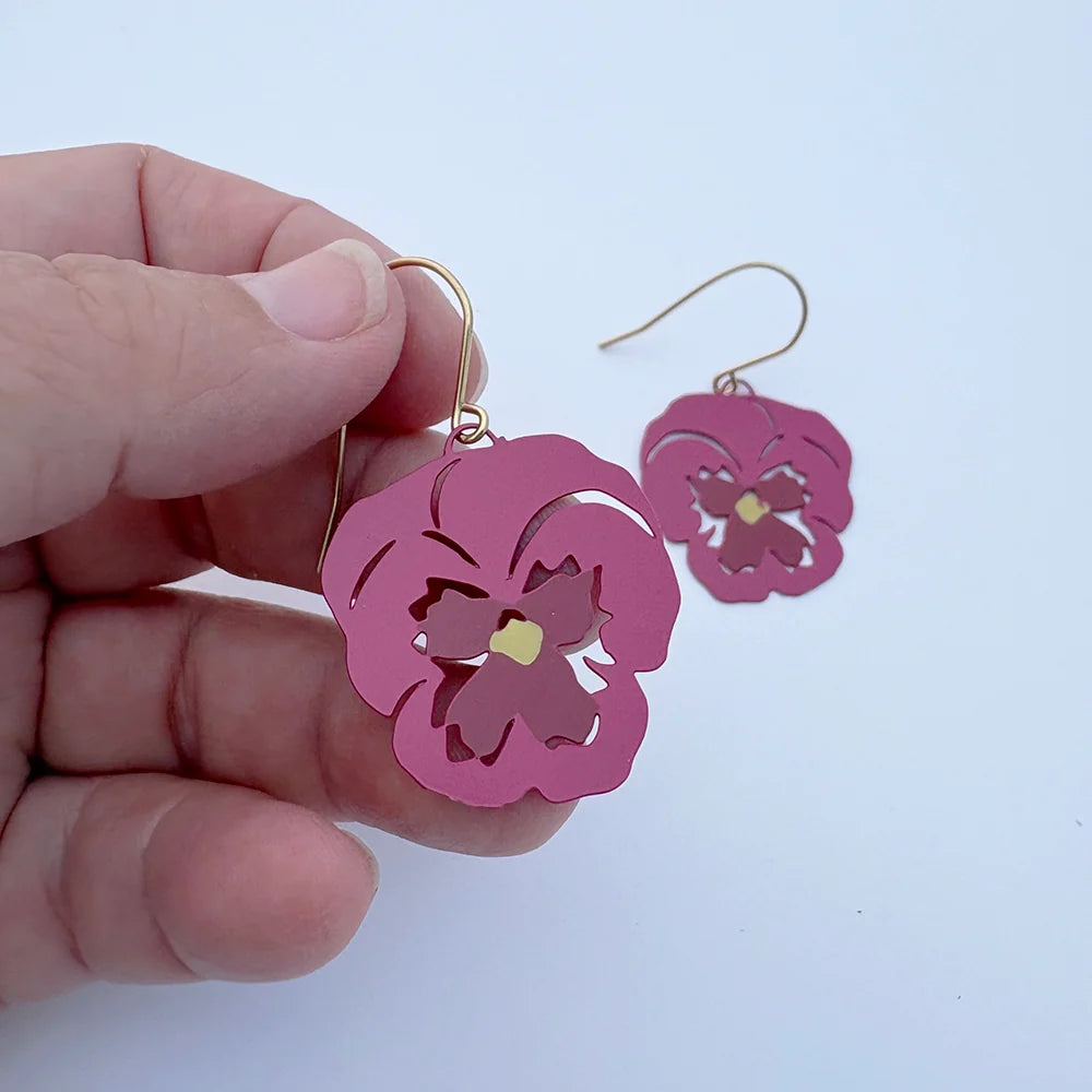 DENZ Pansy Flower Pink earrings held by a hand against a light blue background