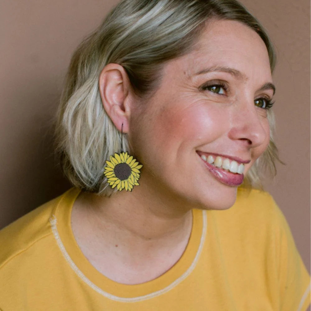 A smiling woman with short blonde hair wearing a DENZ Sunflower Earrings in Brown & Yellow, shown in a close-up portrait against a plain beige wall.
