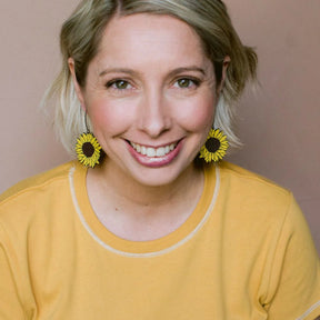 A smiling woman with short blonde hair wearing DENZ Sunflower Earrings in Brown & Yellow and a mustard yellow top, photographed against a plain dusty pink background.