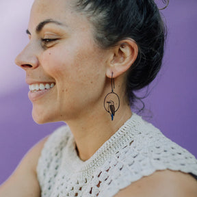A close-up side profile of a smiling woman wearing a DENZ black wire hoop earring with a small bird silhouette and a blue accent, paired with a white crochet top against a solid purple background.