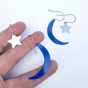 DENZ Moon & Star Stacker Blue crescent moon and silver star drop earrings, with one shown hanging and one held between fingers to demonstrate scale.