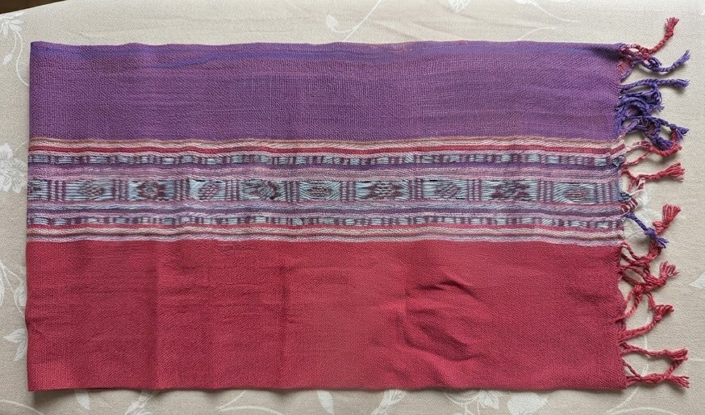 A top-down, flat-lay photograph of a woven textile featuring a rich purple top section, a vibrant raspberry-pink bottom section, and a detailed geometric patterned stripe through the middle. The fabric is finished with twisted fringe tassels along the right edge and laid flat