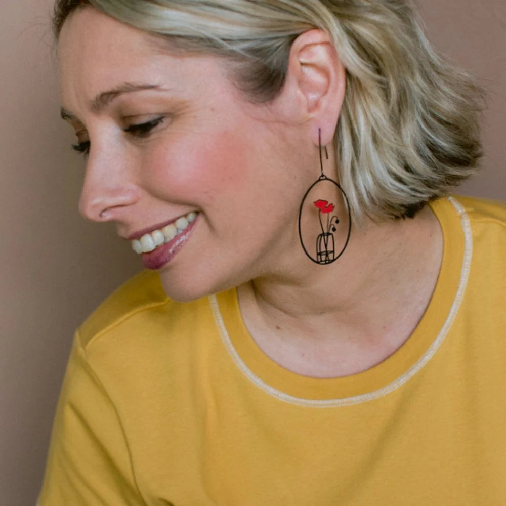 A woman with short blonde hair wearing a yellow shirt and DENZ Poppy Vase earring, shown in a close-up portrait against a plain beige background.