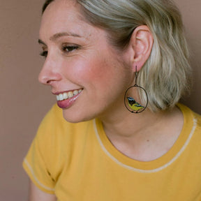 A woman in a yellow t-shirt wearing a DENZ Spotted Pardalote earring with a small yellow and blue bird hanging inside, shown in a close-up profile 
