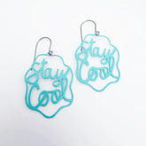 A pair of DENZ mint blue laser-cut metal dangle earrings with the phrase 'Stay Cool' in cursive script.