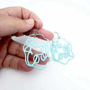 A pair of DENZ mint blue laser-cut metal dangle earrings with the phrase 'Stay Cool' in cursive script, held by a person's hand against a plain white background.