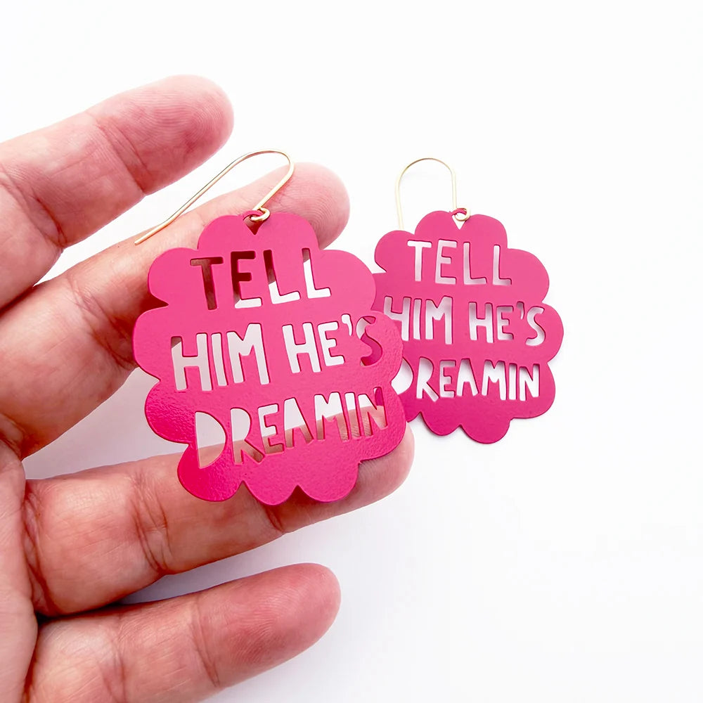 A hand holding a pair of DENZ bright pink scalloped metal earrings with cutout text that reads 'TELL HIM HE'S DREAMIN'. The earrings have gold-tone wire hooks and are shown against a plain white background.