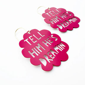A pair of DENZ bright pink metal dangle earrings with scalloped edges and cutout text reading 'TELL HIM HE'S DREAMIN'', shown on a clean white background.