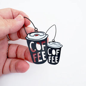Two novelty DENZ Coffee Cup  earrings held by a hand against a plain white background. The earrings are shaped like takeaway coffee cups, with one larger and one smaller, both in black with white lettering that reads 'COFFEE' and light gray lids, hanging from thin black wires.