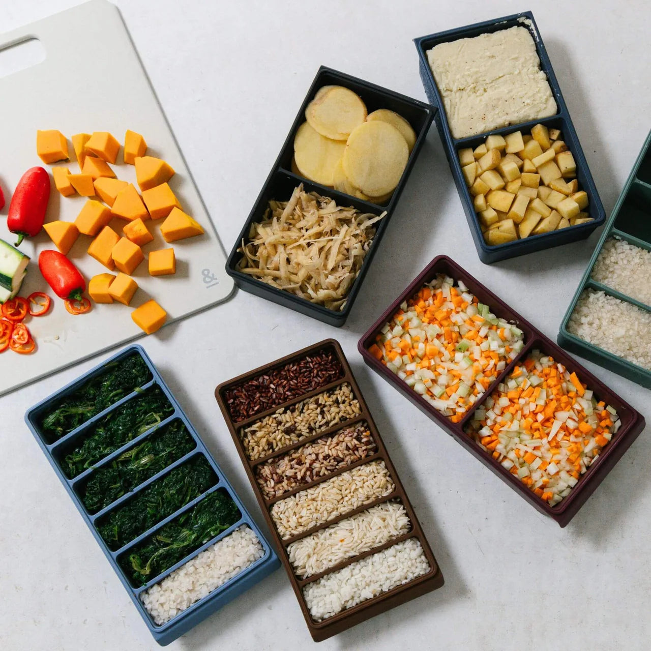 Seed & Sprout silicone freezer trays in assorted 2, 4, and 6-portion sizes filled with prepped vegetables, grains, and potatoes alongside a cutting board.
