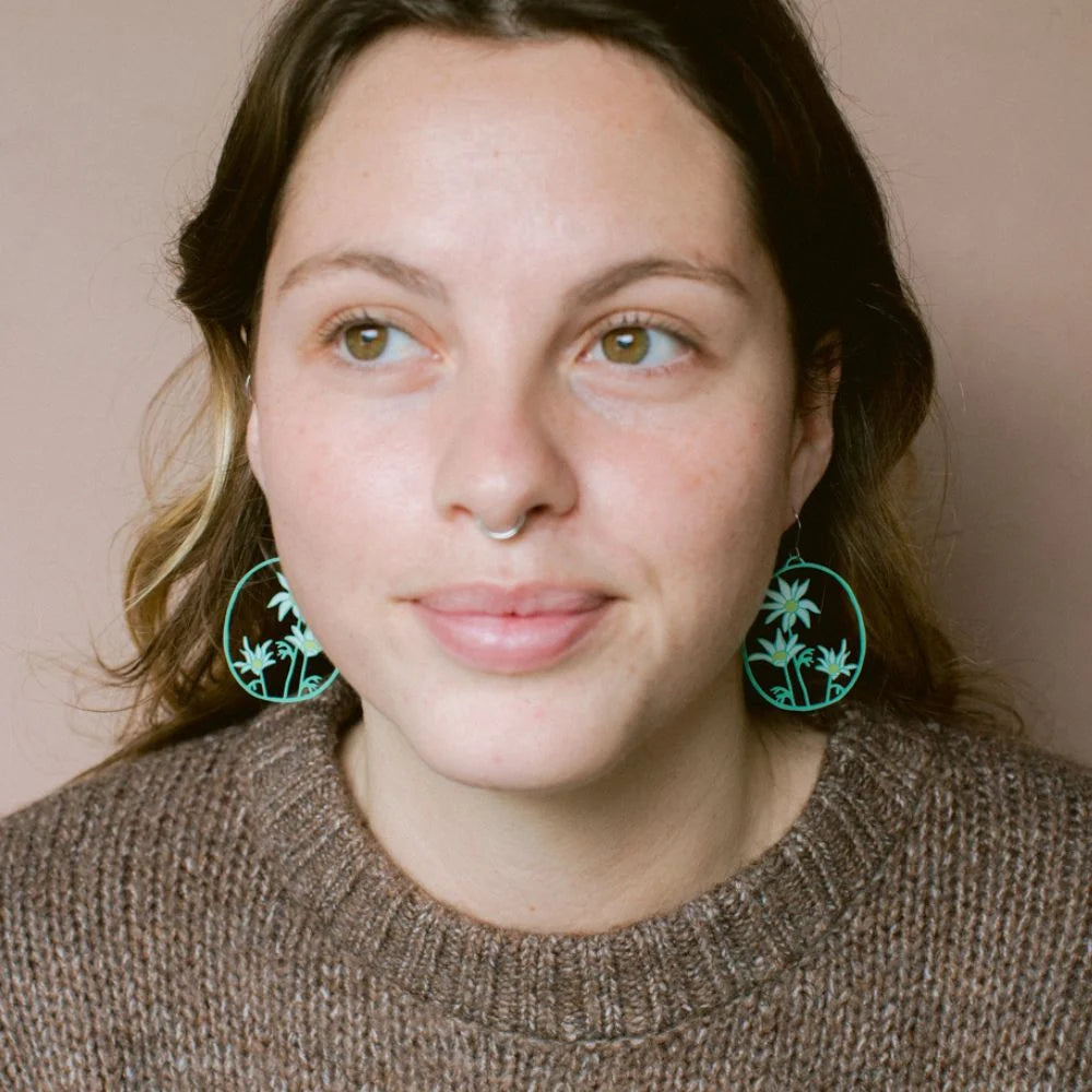 A close-up portrait of a woman wearing DENZ Flannel Flower earring, paired with a brown knit sweater against a solid beige background.