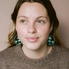 A close-up portrait of a woman wearing DENZ Flannel Flower earring, paired with a brown knit sweater against a solid beige background.