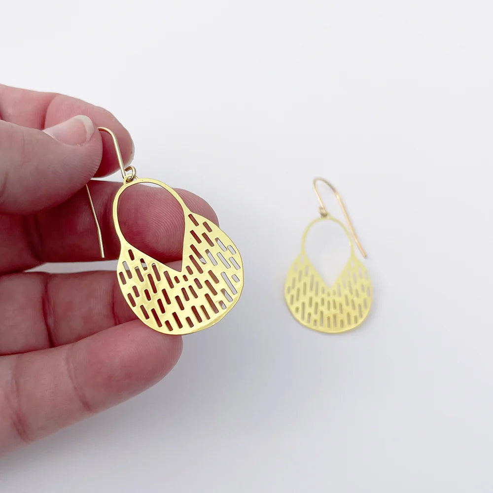 A hand holds a DENZ Callisia gold dangle earring with a geometric cutout pattern, with a second matching earring displayed on a plain white background.