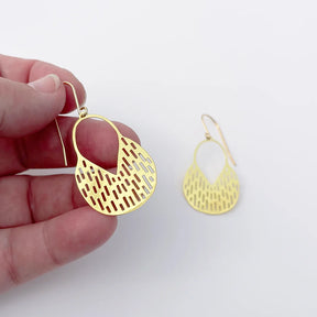 A hand holds a DENZ Callisia gold dangle earring with a geometric cutout pattern, with a second matching earring displayed on a plain white background.
