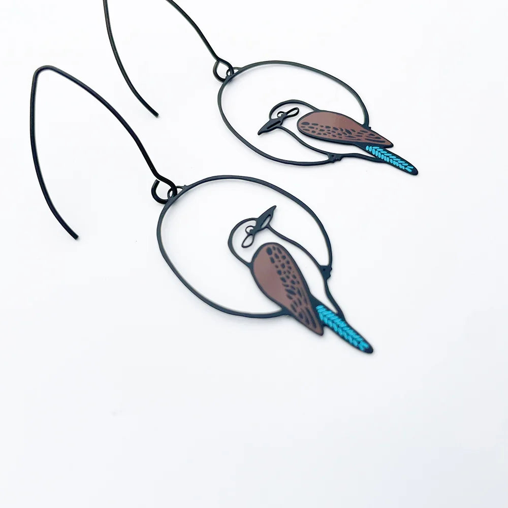 A pair of DENZ black metal wire hoop earrings featuring hand-painted birds with brown speckled bodies and bright turquoise-blue tails, displayed on a clean white background.