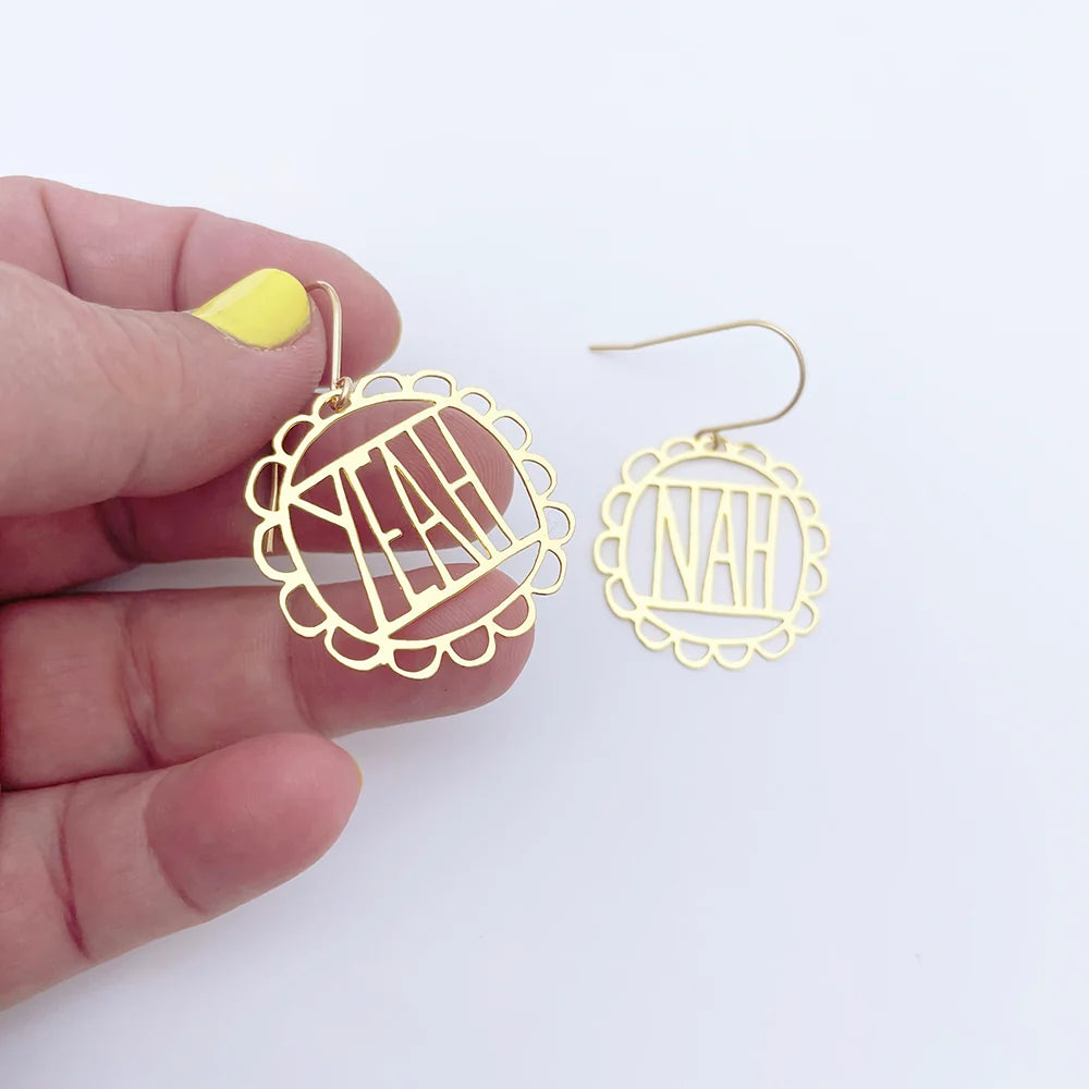 A pair of DENZ gold-toned oval dangle earrings with scalloped edges and cutout lettering "'YEAH NAH", shown on a white background. One earring is held by a hand with yellow nail polish, and the other hangs separately.